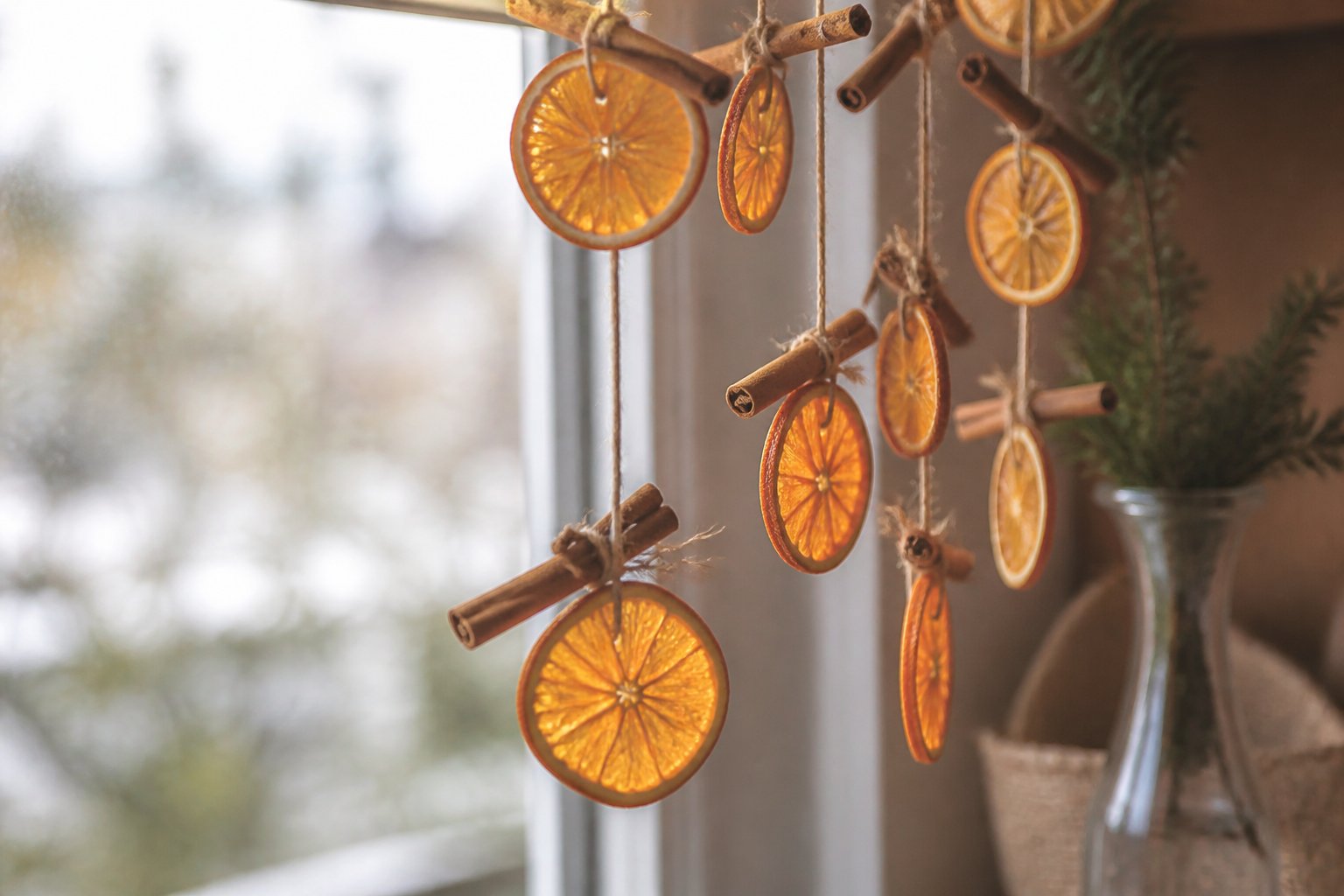 Tranches d’orange séchées enfilées sur une ficelle avec des bâtons de cannelle, devant une fenêtre